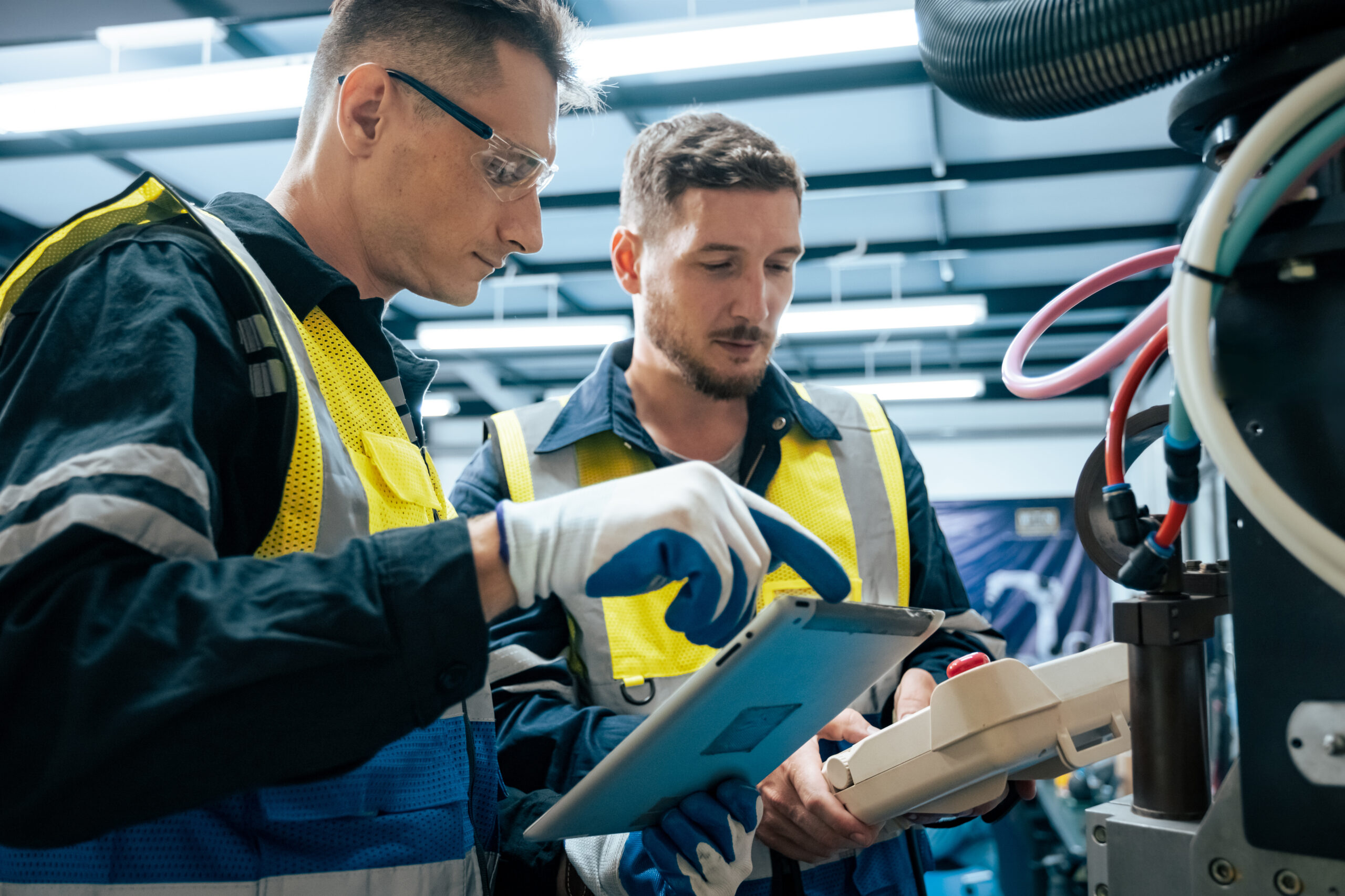 Two male engineers wearing boiler suits and hi-vis vests operate factory machinery in a factory setting under strip lighting